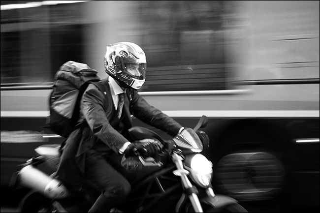 mężczyzna w garniturze jadący na motocyklu z zamazanym autobusem jadącym za nim